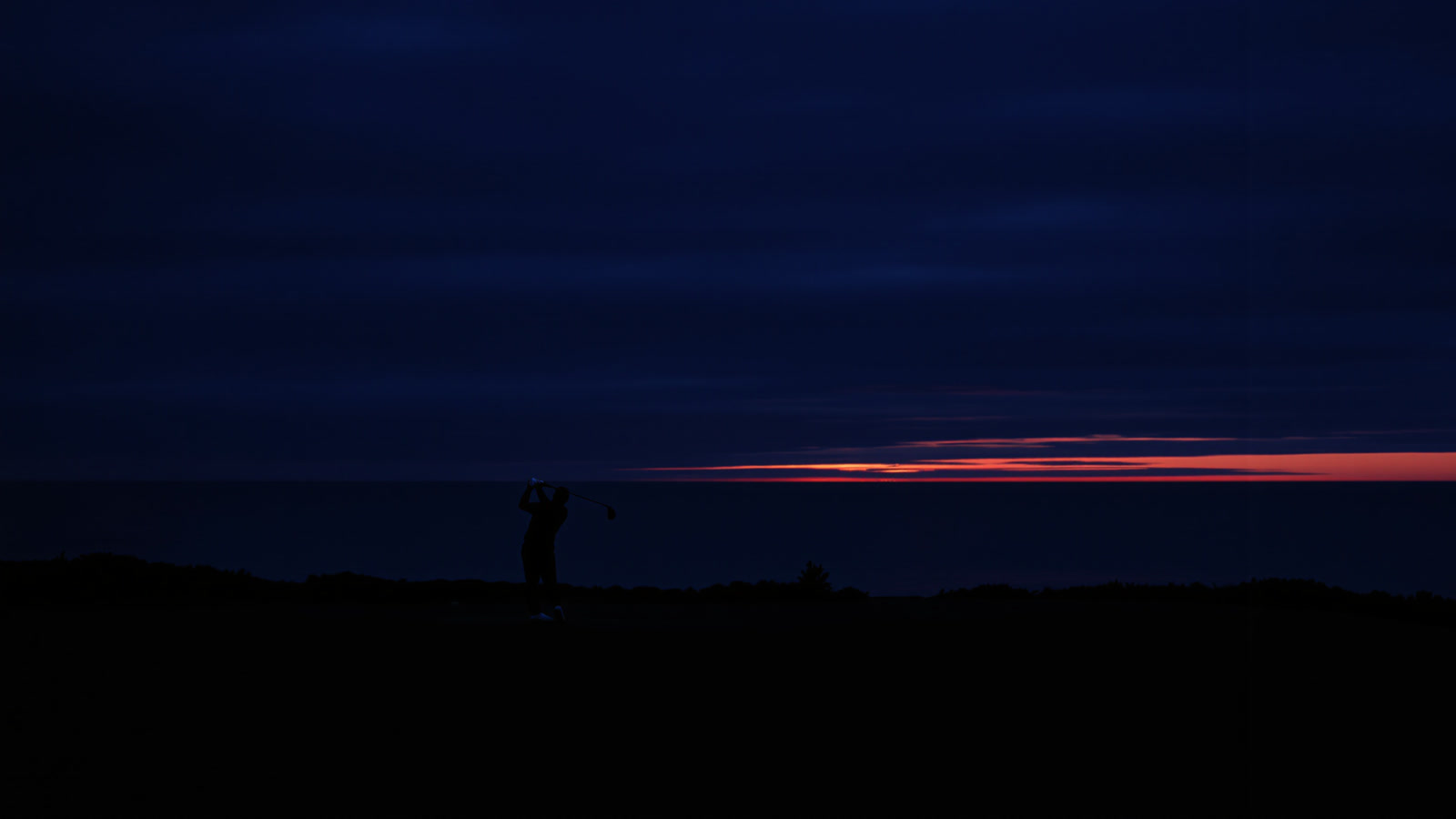 A golfer silhouetted at the top of the backswing at dusk, the last band of light on the sea horizon.