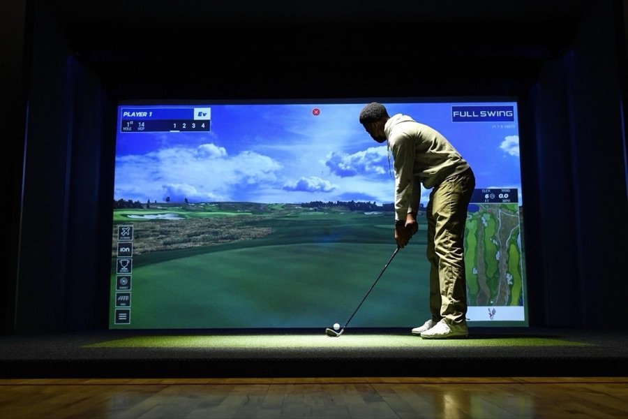 A golfer swinging in front of a large simulator screen in a darkened indoor bay.