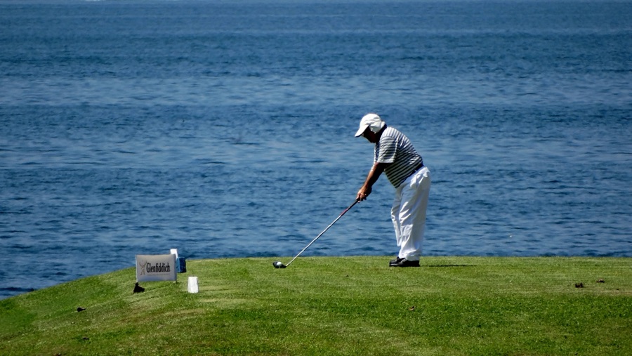 A golfer swinging on a coastal course, the club coming through and the sea visible beyond the fairway.