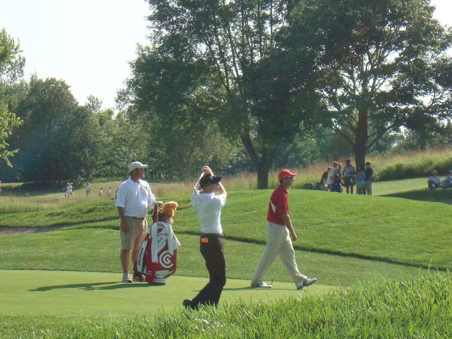 A wide view of a golf hole from behind the tee, players and spectators small in the frame.