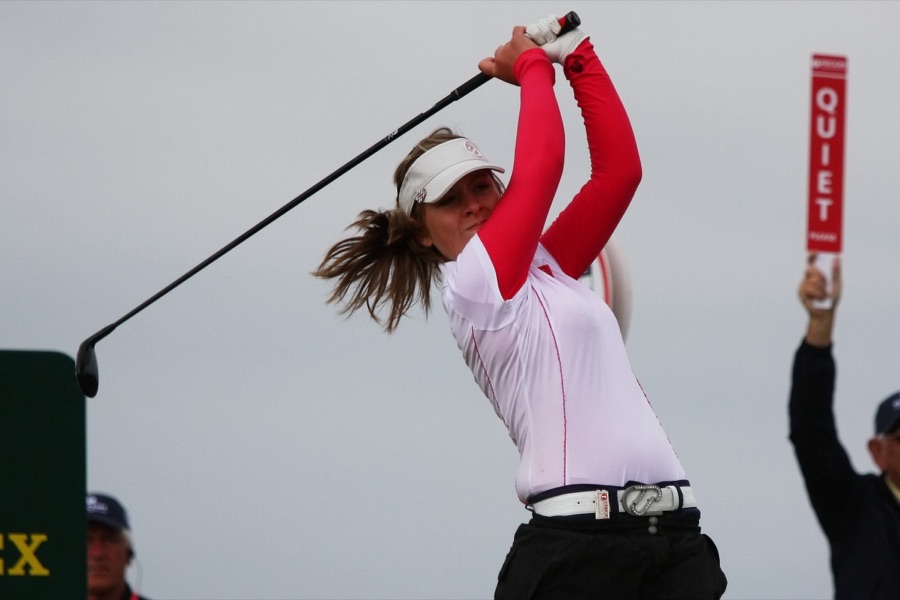 A golfer in a red top at the top of the backswing, both arms up and the club behind the head.