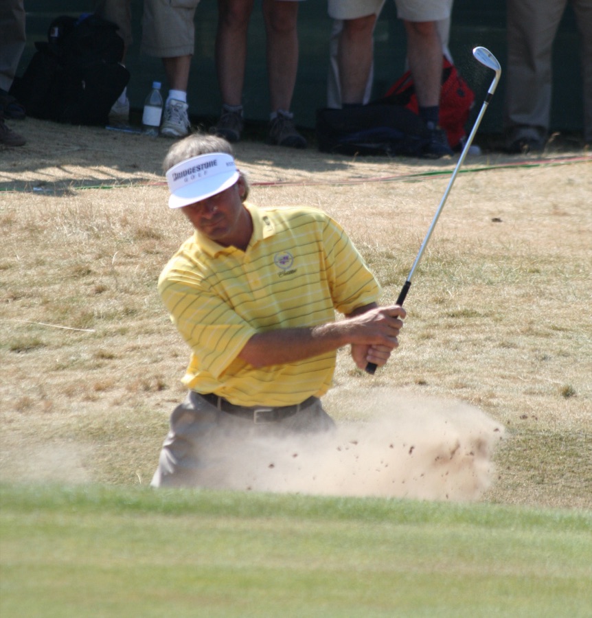 A golfer in a yellow shirt swinging out of a bunker, sand spraying in front of the clubhead.