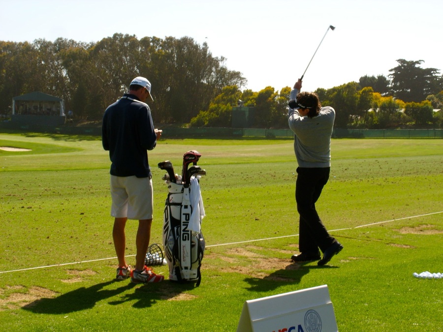 Two golfers on a practice range, one mid-swing and one watching, with bags and clubs between them.