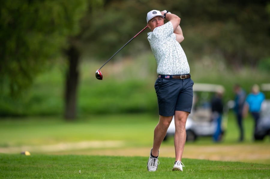 A golfer in shorts and a cap holding the finish on a tree-lined course, spectators behind.