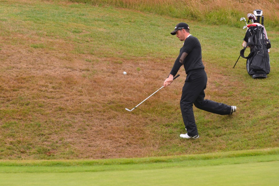 A golfer in dark clothing playing a short shot from the fringe, a golf bag standing on the grass behind.