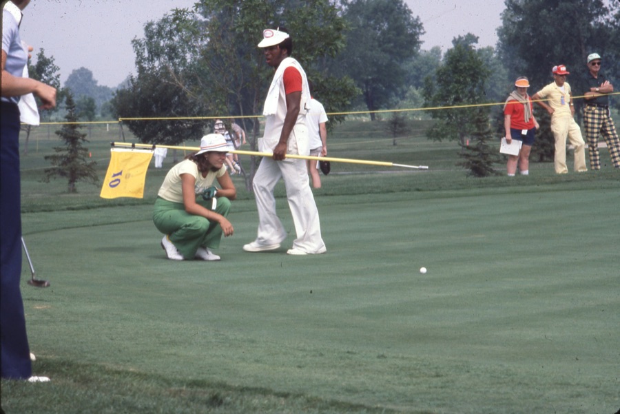 A golfer playing from short rough beside a green while a caddie and spectators watch from behind.