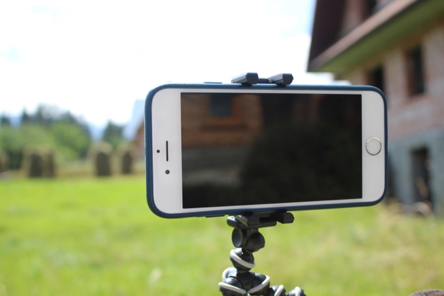 A smartphone mounted on a small tripod on the grass at a driving range, framed on a golfer taking a swing in the background.