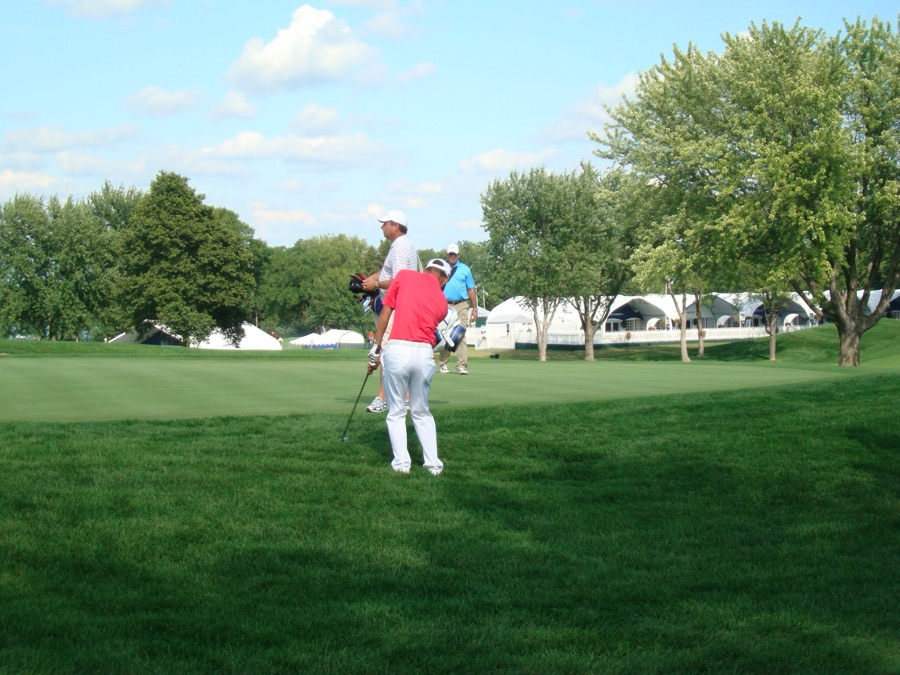 A golfer playing a shot from short grass toward a distant green, photographed from behind at a distance.