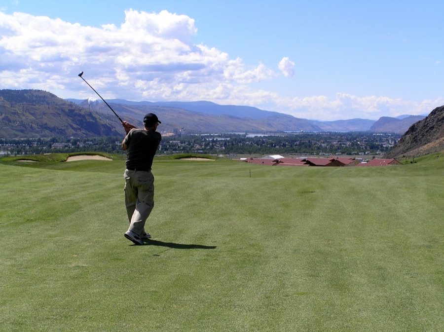 A golfer standing over a ball on a fairway, photographed face-on from a distance with mountains behind.