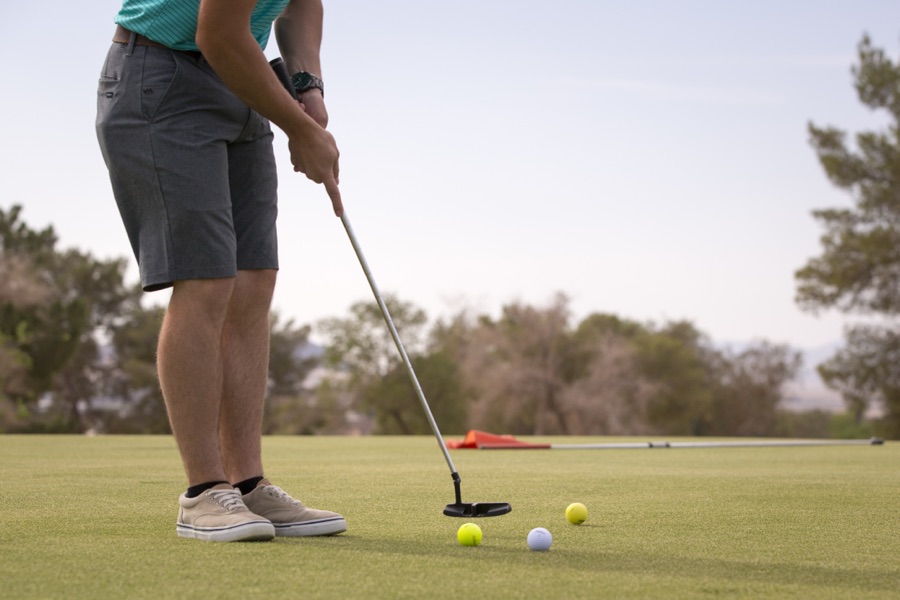 A close view of a golfer's hands and lower body at address, the club soled behind a ball on a mown tee area.