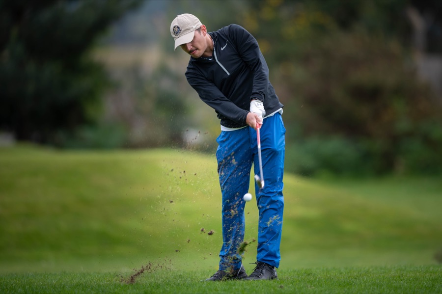 A golfer bent over the ball in a navy top and cap, upper body and club visible against a blurred background.