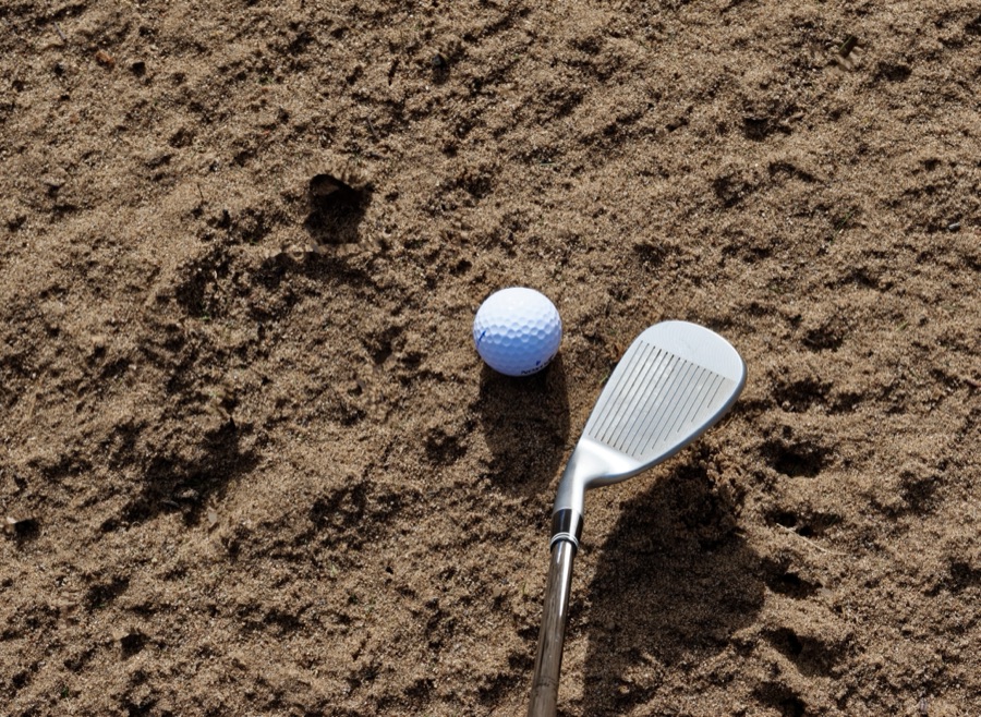 A close view of a wedge resting in sand beside a golf ball, the face and the neck of the club both in frame.