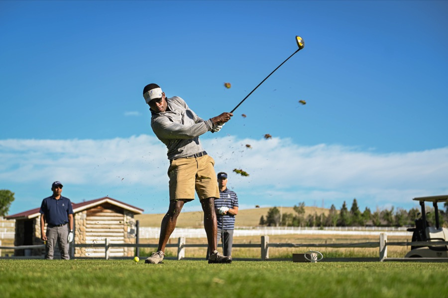 A golfer swinging on a driving range in bright sun, seen from behind and to the side with the tee line and a low fence beyond.