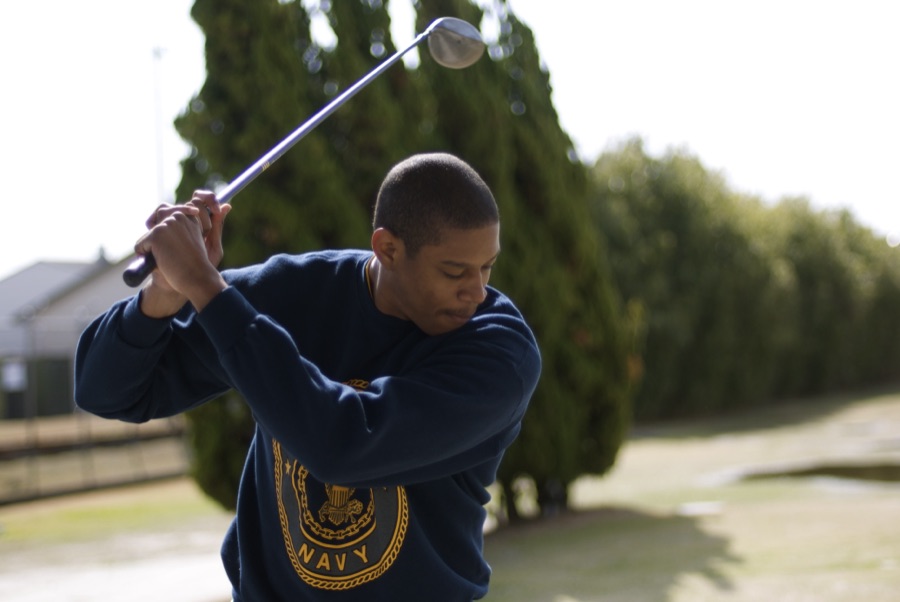 A golfer in a dark sweatshirt at the top of the backswing, photographed close against a hedge.