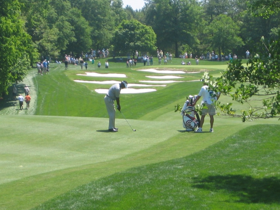 A wide view of a golf hole with players walking the fairway and a bunker in the foreground.