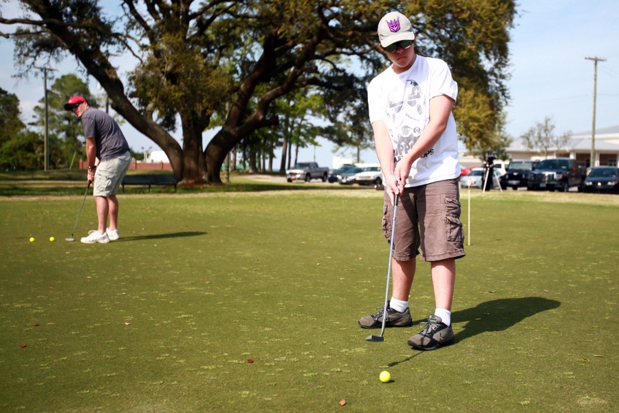 Two golfers putting on a green in bright sun, one standing over the ball and one watching.