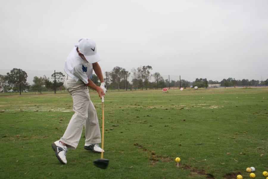 A golfer in fatigues mid-swing on a bare practice ground, scattered balls and a distant tree line beyond.