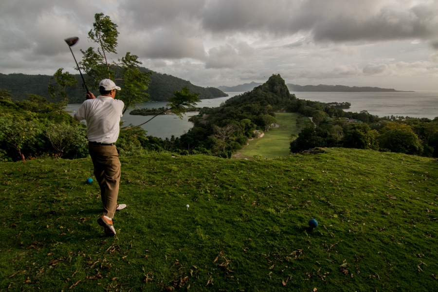 A golfer swinging on a hillside course above a bay, sea and headland in the distance.