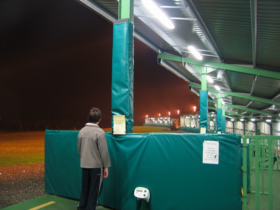 A line of covered bays at a driving range, seen from one end with a golfer at the far stall.