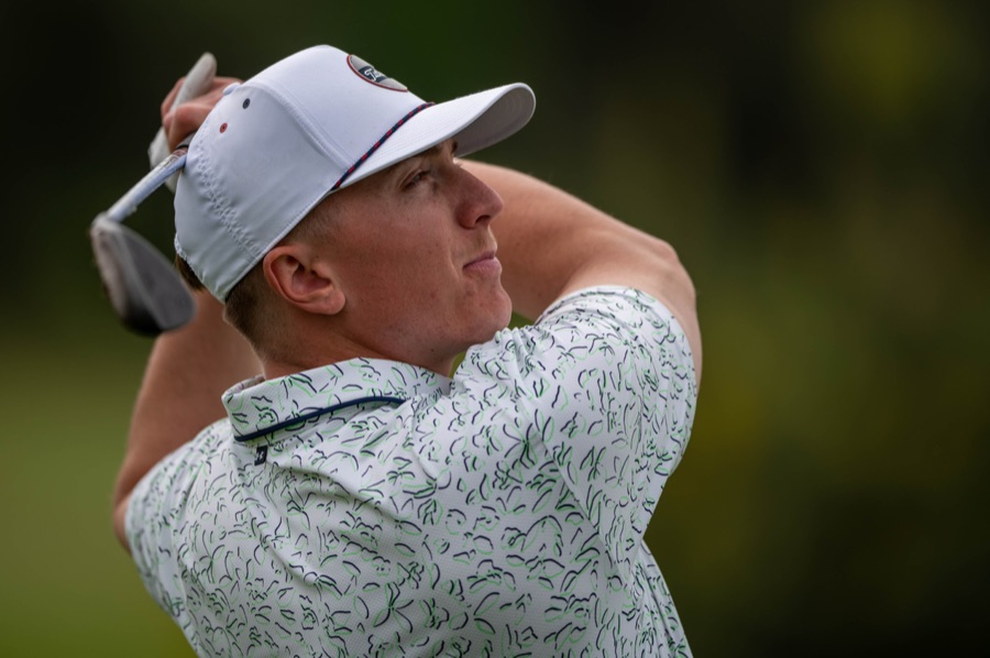 A golfer in a cap and patterned shirt at the top of the backswing, arms and gloved hand filling the frame.