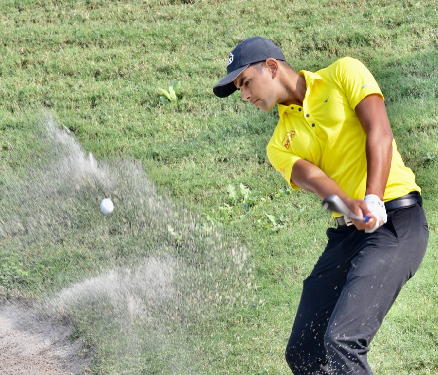 A golfer in a yellow shirt hitting out of a bunker, a cloud of sand thrown forward by the clubhead.