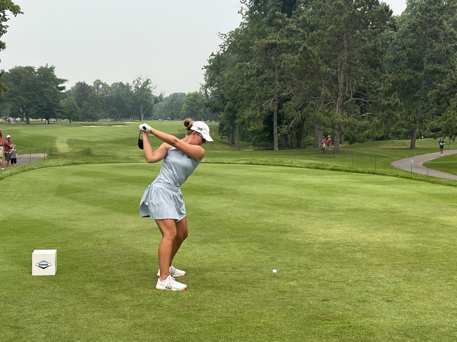 A golfer in a white skirt and cap swinging on a fairway, the club well past the shoulders.