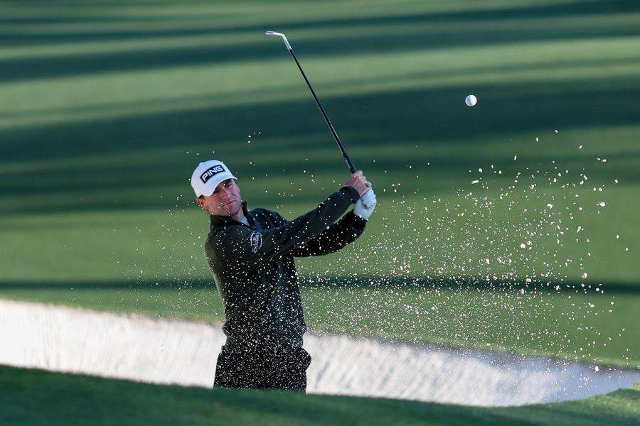 A golfer playing out of a greenside bunker, sand thrown up in front of the clubhead.