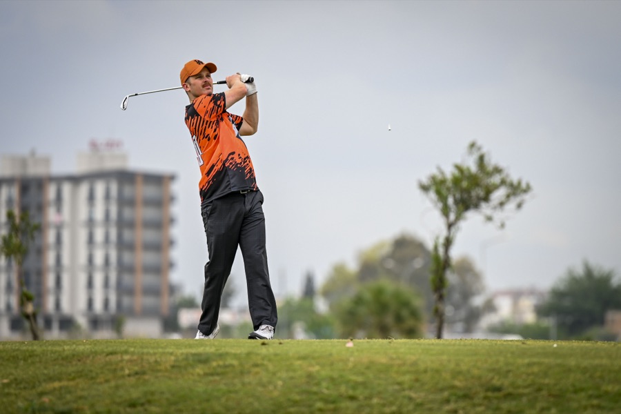 A golfer in an orange shirt mid-swing on a wide, dry course, the whole body in frame from the side.