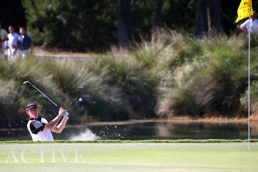 A golfer playing out of a bunker beside a line of tall trees, sand lifting in front of the club.