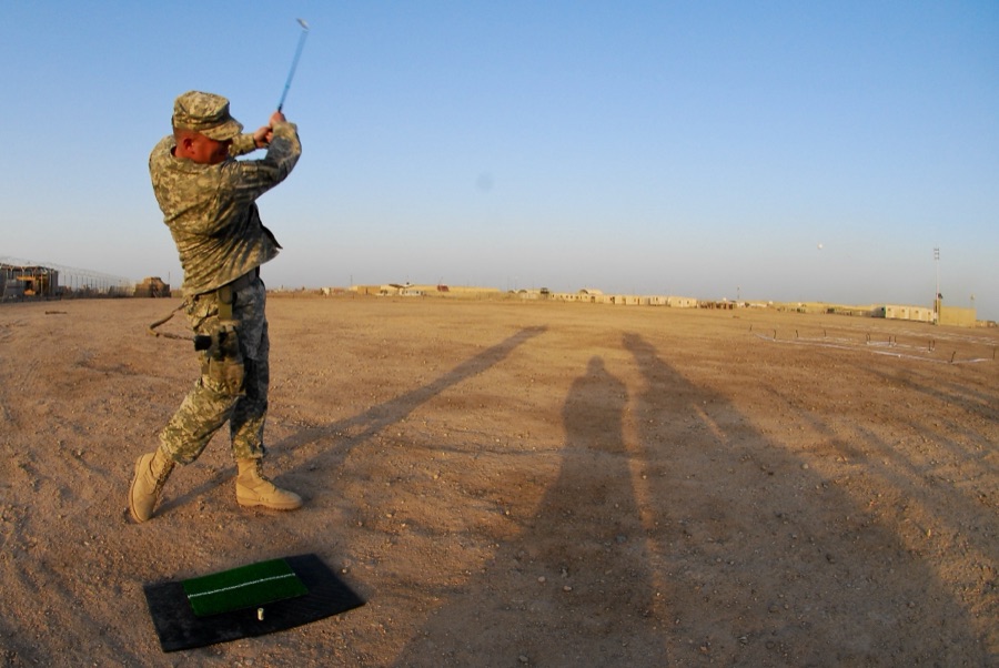 A golfer in fatigues swinging on a dusty practice range, scattered balls on the ground ahead.