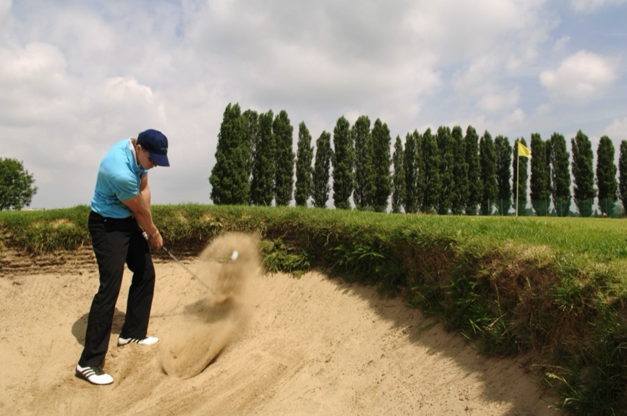 A golfer standing in a deep sand bunker, playing out toward a green lined with tall trees.