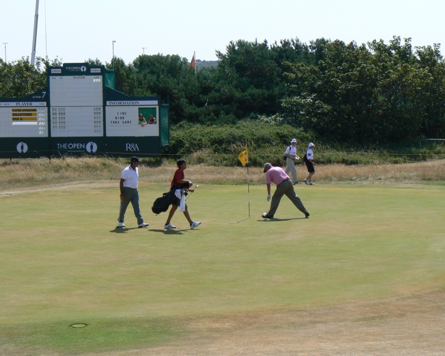 A player crouching to read a putt on the green at the Open Championship, another walking in with a caddie behind.