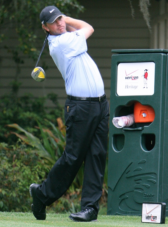 A golfer holding a full finish beside a course bin, back arched and weight fully on the lead foot.