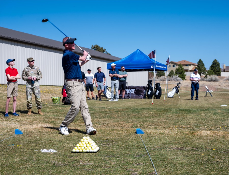 Golfers hitting balls side by side at an outdoor driving range, a marquee and a line of flags behind them.