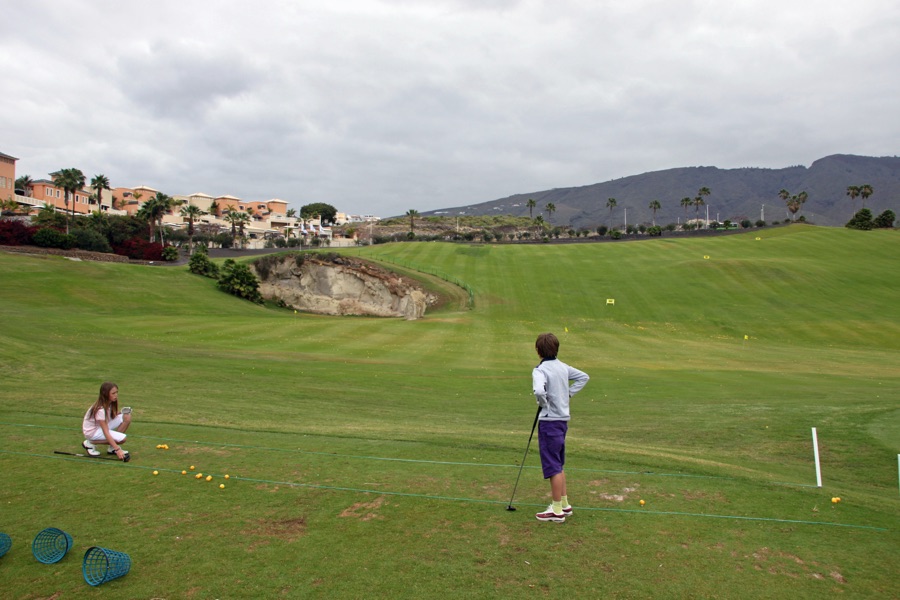 A wide view of a quiet practice range with a single golfer hitting balls, hills in the distance.