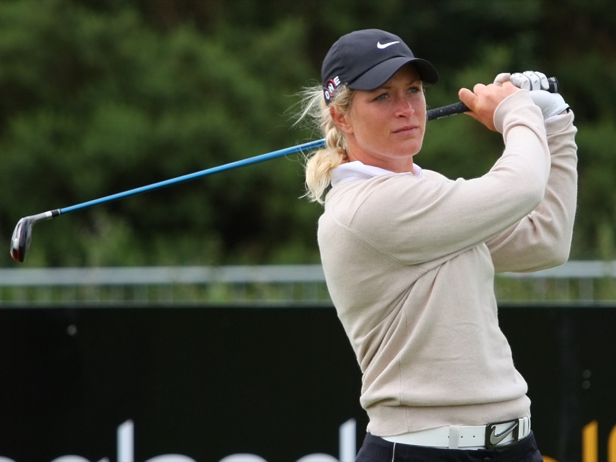 A golfer in a pale sweater at the end of the swing, hands high and body turned through toward the target.